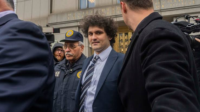 A young man with curly hair, wearing a dark suit and striped tie, exits a building flanked by a U.S. Postal Police officer and other individuals, with a news cameraman capturing the moment. The image relates to the post as it likely depicts Sam Bankman-Fried following the U.S. Senate's unanimous opposition to granting him clemency.