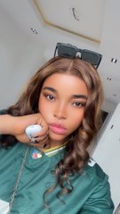 A vertical selfie of a young woman with long brown hair posing in a green top, resting her chin on her hand near a wall socket.