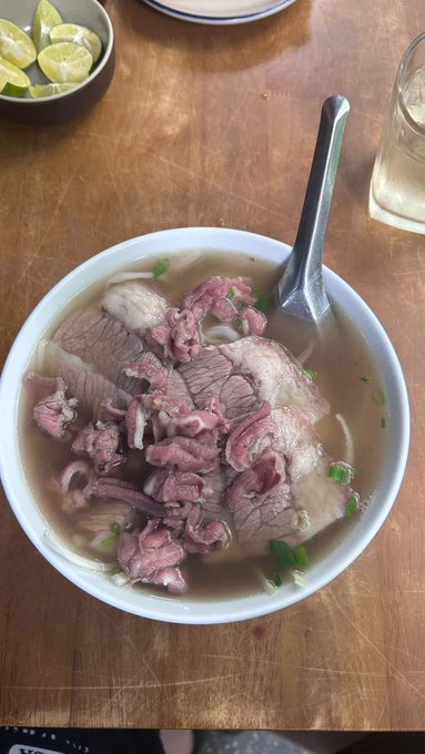 A bowl of Vietnamese pho featuring rare beef slices and meatballs in clear broth, served on a wooden table alongside a small dish of lime wedges.