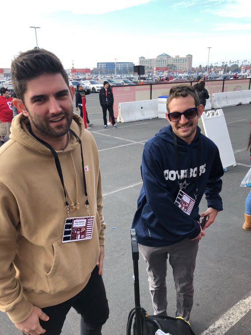 Two smiling young men stand outdoors at what appears to be a parking lot event; one wears a tan Champion hoodie and a lanyard, while the other wears sunglasses and a navy blue 'Cowboys' hoodie with a lanyard.