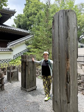 A person wearing a black tank top and patterned pants stands with arms outstretched between two tall, weathered wooden pillars at what appears to be a traditional Japanese temple site.