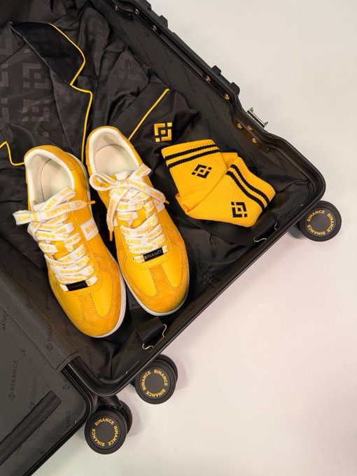 A vibrant yellow sneaker and matching sock set is packed neatly inside an open suitcase with black patterned lining and "BINANCE" branded wheels, perfectly embodying the "Pack a box of good luck!" theme with its bold, travel-ready styling.
