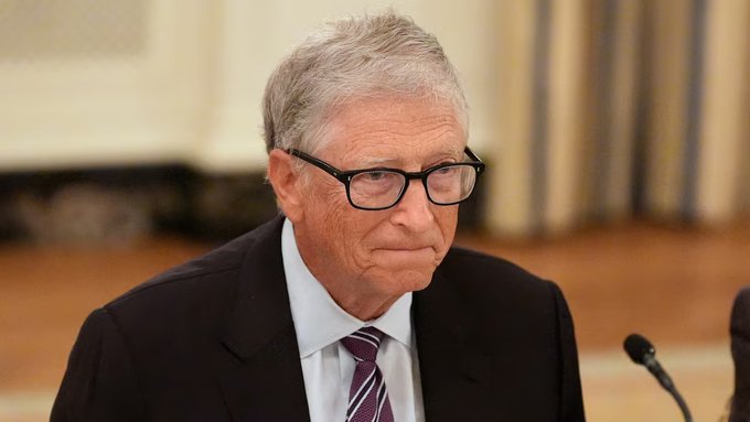 Bill Gates, wearing glasses and a dark suit with a striped tie, sits at a formal setting with microphones visible, appearing serious as he speaks. The image accompanies a post critiquing the Gates Foundation's $1 billion pledge to bridge the AI access gap, suggesting that his foundation's donation-based approach is insufficient compared to tech giants' commercial spending on AI products.