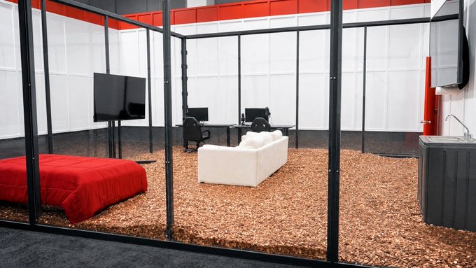 A modern exhibit room enclosed by glass walls features a red bed, white sofa, flat-screen TV, computer monitors, and a sink, all set on a unique cork or wood-chip floor. The image ties to the caption "No one has guessed the correct number yet!" by playfully suggesting this oddly furnished space is not a dwelling but a puzzle—inviting viewers to determine the exact number of items within the room.