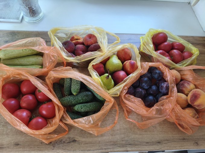 Several plastic grocery bags filled with fresh produce, including tomatoes, cucumbers, peaches, pears, plums, and apricots, are arranged on a wooden table.