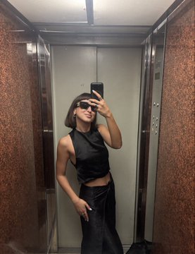 A woman taking a mirror selfie in an elevator, wearing a black sleeveless top, long leather skirt, and sunglasses while holding her phone.