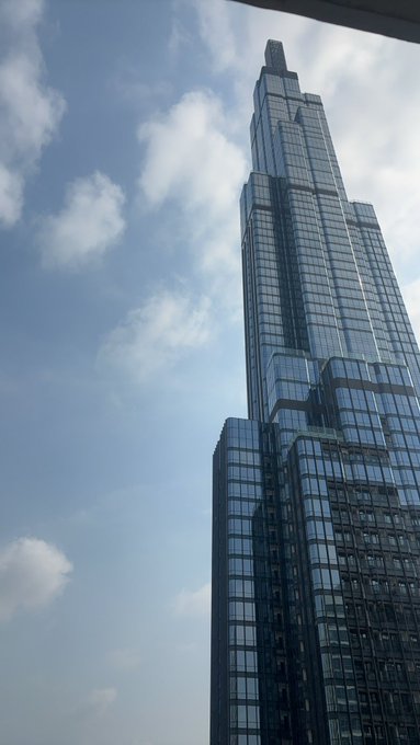 A towering modern skyscraper with a glass facade dominates the right side of the frame, set against a partly cloudy blue sky, suggesting the corporate or commercial environment associated with General Motors. The image appears to reflect GM's presence in an urban business district, possibly highlighting its headquarters or a significant corporate location.