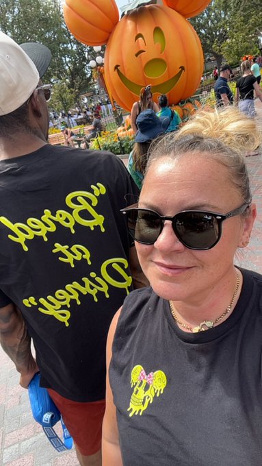 A woman wearing a "Party is Dead" shirt and sunglasses takes a selfie with her companion in front of a giant Mickey Mouse pumpkin display at Disney, capturing a festive Halloween park moment.