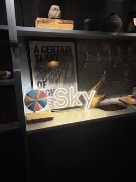 The image features a poster with the word "sky" written on it, which is displayed prominently above a sink area. The sink appears to be made of marble or granite, adding an elegant touch to the scene. Surrounding the sink are various objects such as books and wine glasses, suggesting that this space might be used for both work and relaxation. A small bird statue can also be seen in the image, placed on a shelf above the sink area. The overall atmosphere of the room seems to be one of sophistication and tranquility.