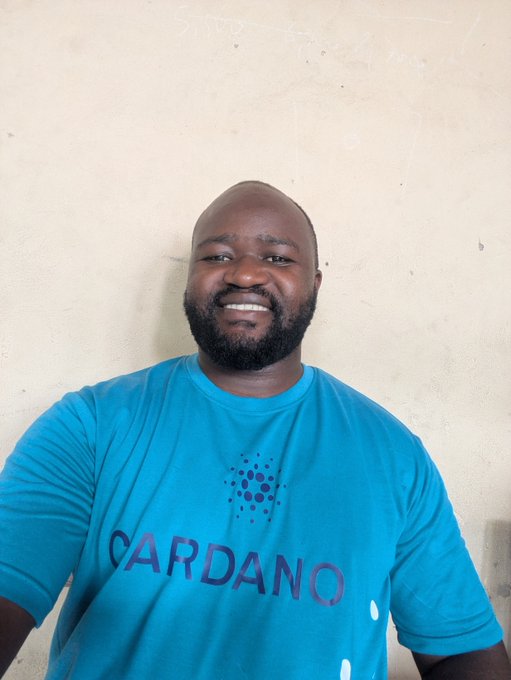 Smiling bald man with a beard wearing a blue Cardano t-shirt against a plain wall.