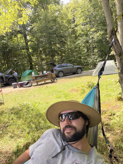 A selfie of a bearded man wearing a wide-brimmed hat and sunglasses relaxing in a hammock at a wooded campsite, with tents, vehicles, and a picnic table visible in the background.