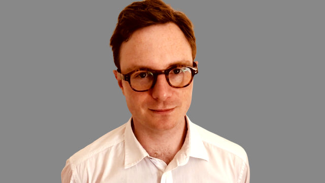 A portrait photograph of a man with short reddish-brown hair and glasses, wearing a white collared shirt against a plain gray background.