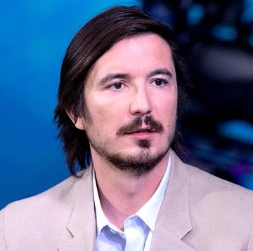 A man with long dark hair, a mustache, and goatee, wearing a beige blazer over a white shirt, appears on what looks like a news or broadcast set with a blue background. The image likely depicts a financial analyst or commentator discussing the breaking news about two Robinhood ($HOOD) employees being charged with fraud for alleged insider trading on Hyperliquid.