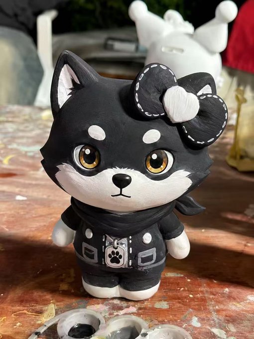 The image features a black and white cat figurine with a bow around its neck, sitting on a table covered in paint splatters. The cat appears to be wearing a jacket and has a name tag attached to its collar that reads "mame". In the background, there is another cat figurine, which also has a name tag reading "mame".