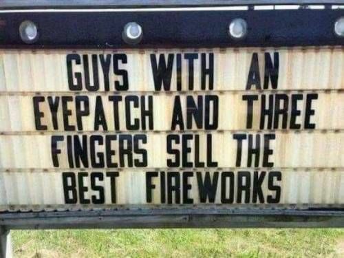 A weathered outdoor marquee sign with black lettering reads "GUYS WITH AN EYEPATCH AND THREE FINGERS SELL THE BEST FIREWORKS." The sign is mounted with visible light bulbs along the top edge, set against a backdrop of green grass.