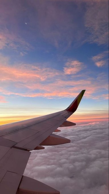 An airplane wing extends over a sea of clouds during a vibrant sunset, with a crescent moon visible in the sky above. The serene aerial view complements the "Get Money" theme, evoking the aspirational journey toward financial success and reaching new heights.