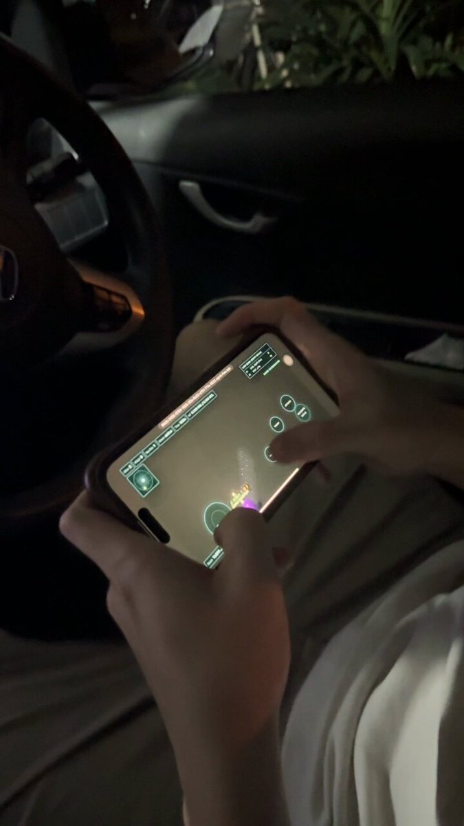 The image shows a person sitting inside a car, holding up their phone displaying an app that appears to be a mobile game or testing platform for smartphones. The person seems to be playing the game while seated in the vehicle. The scene also includes a car's steering wheel and a potted plant nearby, adding to the overall atmosphere of the image.