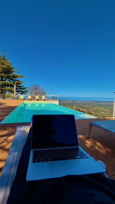 A laptop rests on someone's lap beside a bright turquoise infinity pool overlooking a scenic landscape under a clear blue sky.