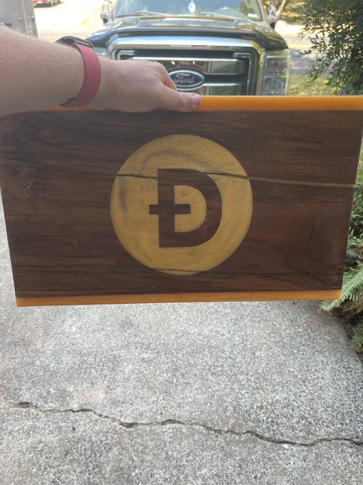 A hand wearing a pink Apple Watch band holds up a wooden plaque featuring a carved Dogecoin 'Ð' logo, with a black Ford truck visible in the background.