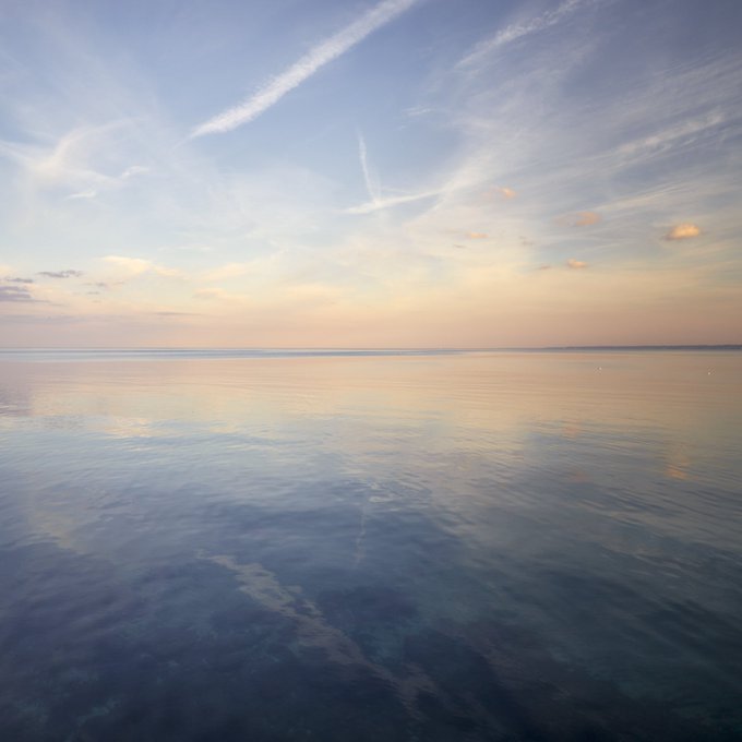 A serene photograph of a calm ocean at sunset or sunrise, with pastel-colored clouds reflected in the still water and a contrail visible in the sky.