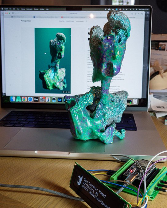 A holographic-style sculpture with iridescent green and purple coloring sits in front of a laptop displaying its image on what appears to be a gallery website, accompanied by a digital message board reading "The Ocean's Pearl! Art is Fast! 😊" with a reference number. The setup reflects a "Sunday Gm" vibe — a creative, art-focused greeting showcasing a finished piece alongside its digital references and maker notes, perfect for sharing Sunday artistic inspiration in a crypto or NFT community context.