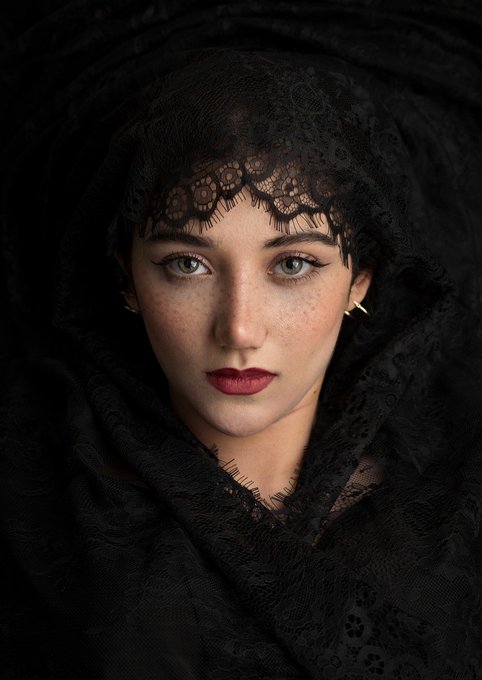 A portrait of a woman with freckles, blue eyes, and bold red lipstick, wearing a black lace veil that drapes over her head and shoulders.