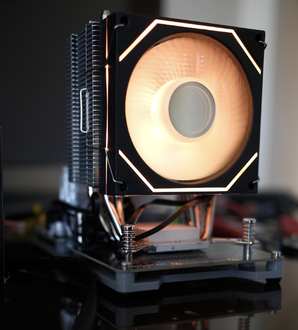 A close-up of a CPU air cooler with an illuminated orange fan, mounted on a motherboard with visible CPU socket screws.