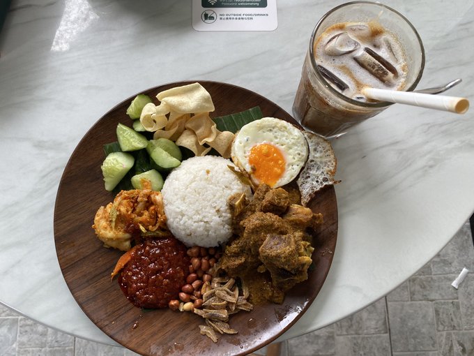 A wooden plate of Nasi Lemak featuring rice, sambal, peanuts, a fried egg, cucumber, and curry sits on a marble table alongside an iced coffee; a partially legible note reads 'NO HOT FOOD' and 'FOOD IS FOR CUSTOMERS, PLEASE KEEP TABLE CLEAN'.