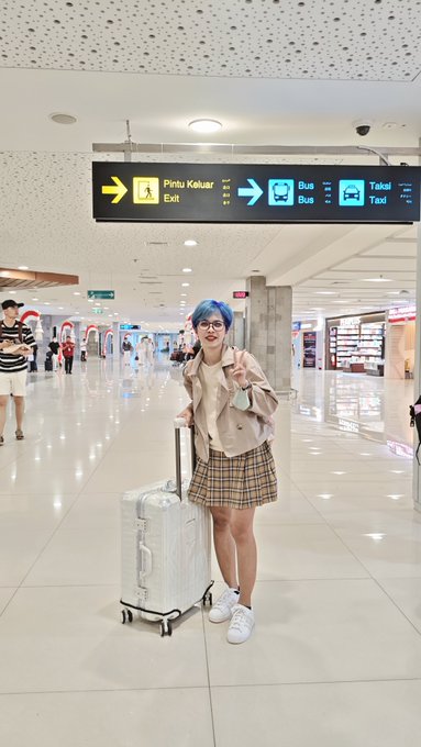 A cosplayer with blue hair wearing a beige vest and plaid skirt poses with a white suitcase in an airport terminal under signs pointing to Exit, Bus, and Taxi.