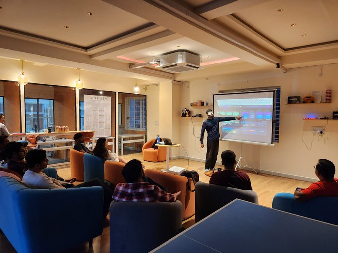 A presenter wearing a mask stands before a projection displaying charts in a modern room with comfortable seating, where several audience members are watching; a poster reading 'BITCOIN' is visible on the wall.