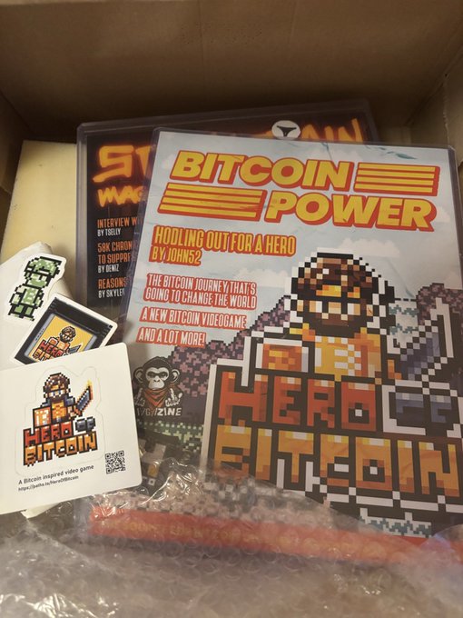 A 'Hero of Bitcoin' merchandise package inside a box, including a 'BITCOIN POWER' magazine, stickers, playing cards, and a pixel art keychain.