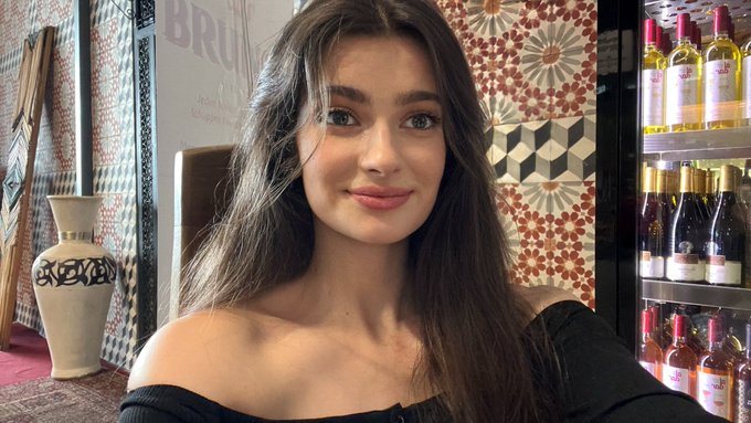 A young woman with long brown hair takes a selfie in what appears to be a shop, with decorative tiles and shelves of bottles visible in the background.