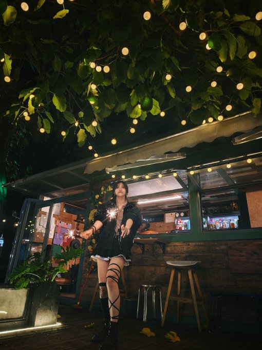 A nighttime photo of a young woman holding sparklers in front of a food truck or kiosk decorated with string lights hanging from a tree.