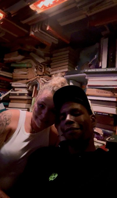 A dimly lit selfie of two people in what appears to be a book-filled room or bookstore, with stacks of books piled high behind them.