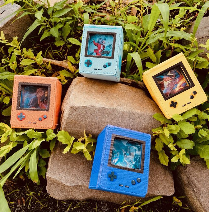 Four colorful Game Boy-style card holders (orange, light blue, yellow, and blue) with game artwork displayed, arranged on rocks among green plants.