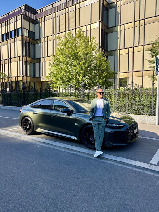 A man in a mint green suit poses beside a matte olive-green Audi RS sedan parked on a city street, with modern glass-fronted buildings in the background. The sleek styling of both the car and his outfit embodies a polished, luxury lifestyle fitting for a "GM Fam" post.