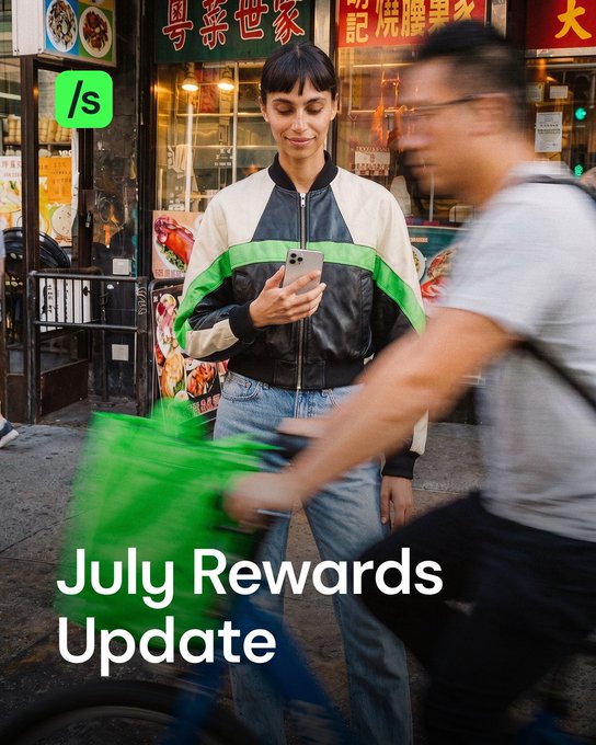 A woman in a green, black, and white jacket looks at her smartphone on a city street, with a man on a green bike riding past, while text reads "July Rewards Update".