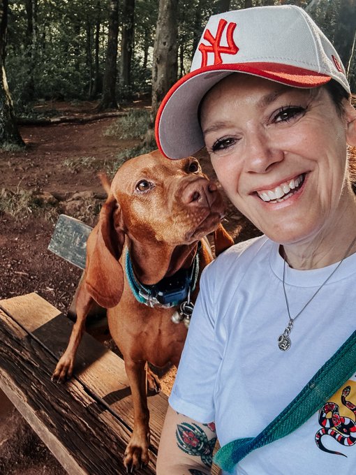 A smiling woman wearing a New York Yankees cap takes a selfie with a brown dog that is licking her cheek in a wooded area.