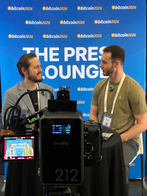 Two men are interviewed in "The Press Lounge" at the Bitcoin 2026 conference, with a small monitor on a camera labeled "212" displaying the live feed against a backdrop featuring repeated "bitcoin 2026" logos—visually capturing the event where the quoted insight about Lightning as a foreign exchange layer for global currencies was shared.
