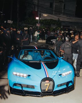 A bright blue Bugatti sports car with French flag stripes on the hood is parked at night while a crowd of people gathers around it.