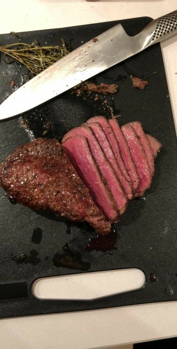 Slices of medium-rare steak rest on a black cutting board next to a large knife and some herbs.