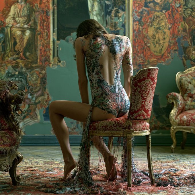 An artistic photograph shows a woman seated on an ornate red floral chair with her back to the camera, wearing a sheer, colorful beaded dress in a room with elaborate wall panels and patterned rugs.