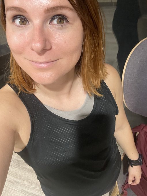 A close-up selfie of a woman with red hair wearing a black athletic tank top over a gray shirt, smiling at the camera in an indoor setting.