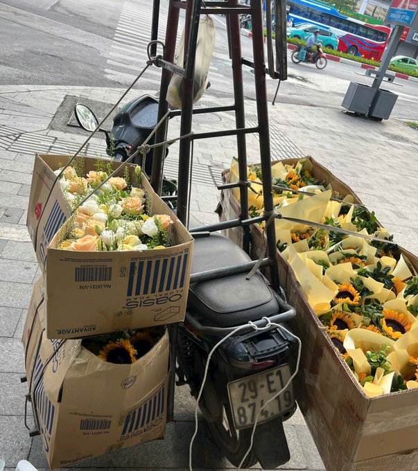 A motorcycle parked on a sidewalk loaded with cardboard boxes containing wrapped bouquets of roses and sunflowers, with a license plate reading '29-E1 874.19'.