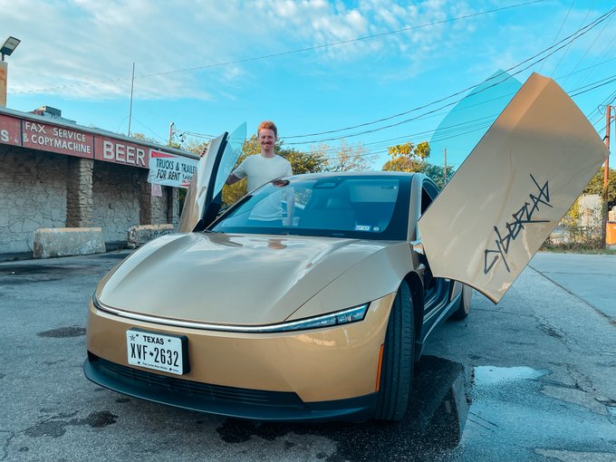 A man standing next to a tan/champagne colored futuristic car with upward-opening scissor doors that have graffiti tags on them, parked outside a building with signs for 'BEER' and 'FAX SERVICE & COPY MACHINE'. The Texas license plate reads 'XVF-2632'.