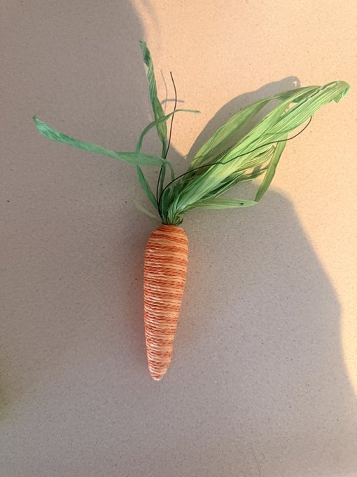 A decorative craft carrot made from orange yarn or string with green paper or fabric leaves, mounted on a plain beige wall.