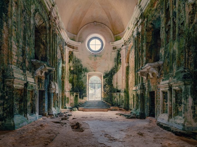 An atmospheric photograph of an abandoned church interior featuring vaulted ceilings, a circular window above, and an arched doorway at the back. The walls and columns are heavily weathered with green moss and peeling plaster.