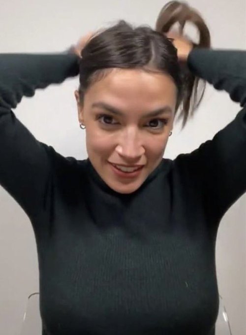A selfie of a smiling young woman with dark hair tying it up, wearing a black ribbed long-sleeve top.