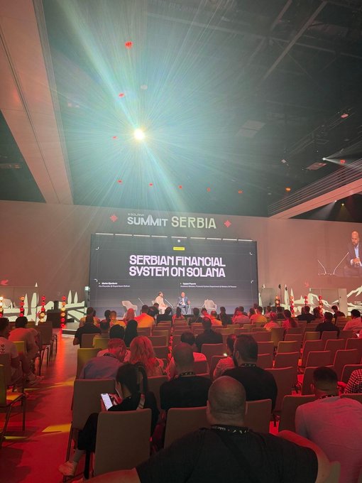 An audience sits in a conference hall attending the 'Solana Summit Serbia,' with a large screen displaying 'SERBIAN FINANCIAL SYSTEM ON SOLANA' under bright stage lights.