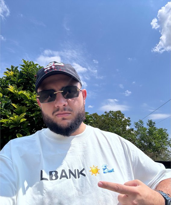 A bearded man wearing sunglasses and a black cap poses outdoors in front of a blue sky, wearing a white t-shirt with 'L8ANK' and sun/cloud emojis while pointing at the text.